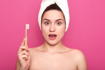 Obraz na płótnie Canvas Image of surprised lady with toothbrush in hand, posing in bathroom while brushing teeth in morning, poses with bare shoulders and white towel on hand, looks shocked, isolated over pink background.