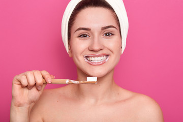 Close up portrait of attractive caucasian smiling dark haired woman isolated on rose studio, posing with towel on head and bare shoulders, has perfect skin, looks at camera with tooth brush in hand.