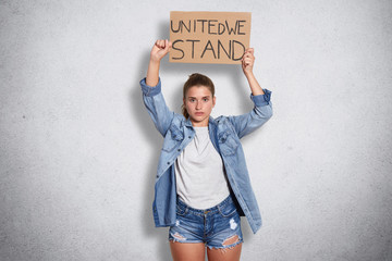 Confident powerful brunette female wearing jeans jacket, shorts, casual white t shirt, holding feminist inscription above her head, having serious facial expression, looking for like minded people.