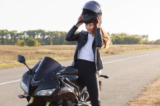 Image of beautiful slender young risk taker sitting on her motorcycle, having trip on her vehicle, fond of extreme activities, wearing helmet off, standing at side of road. Motorcycle tourism concept.