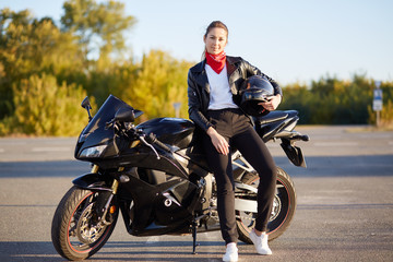Photo of attractive slim female with ponitail, standing near black fast motobike with helmet in hand, wearing stylish outfit, has confident facial expression, poses outdoor. Motorcycle tourism concept