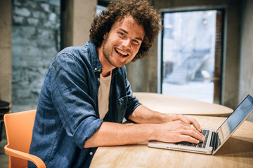 Portrait of happy young man with curly hair using laptop for working and browsing online. Smart Caucasian male reading news, typing on his laptop computer in the modern office. Business and people