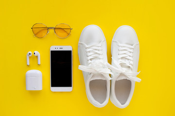 White sneackers, Pink transparent sunglasses, wireless headphones and smartphone are lying on a yellow background.