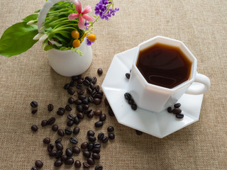 Cup of coffee and coffee beans are next to a vase of flowers on a background sack.