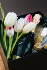 Beautiful awesome black giftbox with wonderfull tulips, bottle of champagne and handmade chocolate candies for celebration on wooden table with dark background