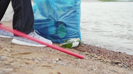Closeup tilt up shot of young beach cleaner with trash picker and bag picking up glass bottle