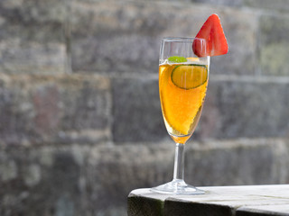 Close up of a champagne flute glass with a fruity punch drink containing a piece of orange and cucumber decorated with a strawberry on the rim of the glass.Copy space and wall in background - Image