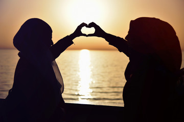 Silhouette of two muslim women  in hijab is standing on the passenger ferry's deck and enjoying sunshine from the ship. Drawing hearts with their hands on the ship at sunset.