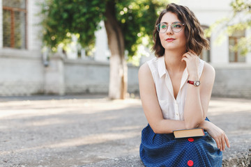 Fototapeta premium Beautiful young brunette woman with short wavy hair, wearing white sleeveless shirt, long blue skirt and glasses reading a book outdoor on old city street. Student, retro outfit.