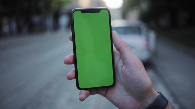 Man's hand shows green screen mock up smartphone display in vertical position in the city street on blurred background.