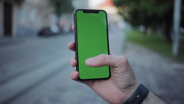 Close-up of guy holding a smartphone mockup in his hand in the city center. Man using a smartphone typing on green screen on blurred background.