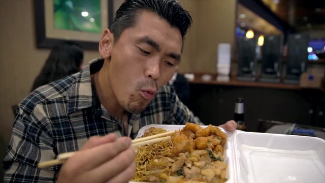 This video shows a hungry young tattooed asian man eating and devouring chinese takeout food straight out of the box with chop sticks.