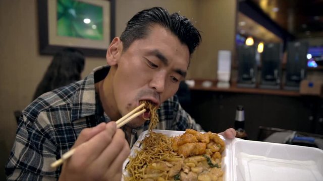 This slow motion video shows a hungry young tattooed asian man eating and devouring chinese takeout food straight out of the box with chop sticks.