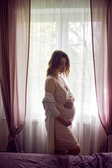pregnant girl in a pink silk nightie stands against the background of a large window with curtains at home