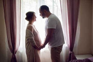 Fototapeta premium pregnant girl in a nightie with her husband in a white t-shirt stands against the window of the house