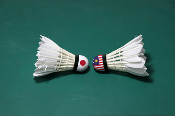 Two used shuttlecocks on green floor of Badminton court with both head each other. One head painted with Japan flag and one head painted with the Malaysia flag.
