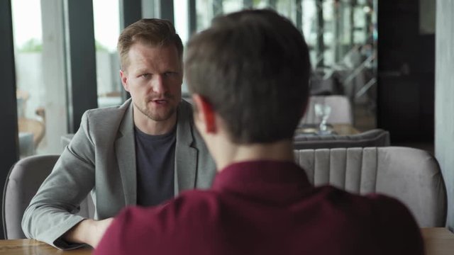 Tracking left shot of two men having business meeting over coffee in cafe. Middle adult businessman with beard sitting at table and explaining points to unrecognizable partner