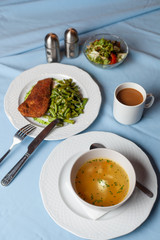 Three course substantial set meal on a blue tablecloth: hot chicken noodle soup, chicken schnitzel with green beans green broccoli salad and a cup of coffee