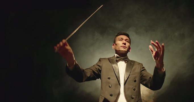 Asian symphony orchestra conductor wearing suit is directing musicians with movement of baton, isolated on black smokey background 4k footage