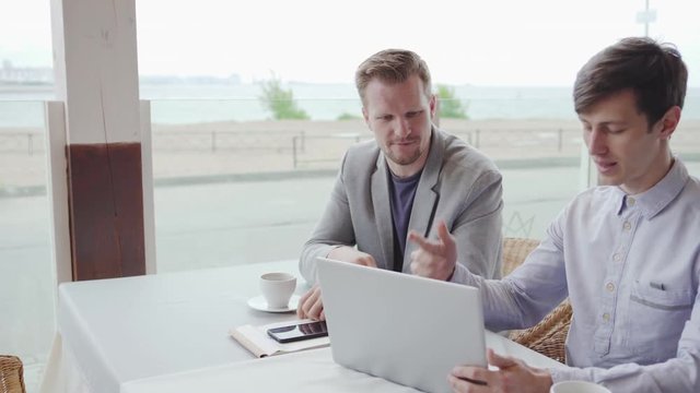 Tracking right shot of two businessmen sitting at table in cafe and discussing project on laptop screen, side view.