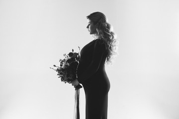 Portrait of young pregnant woman with big flower in her hand. Beautiful model posing in dark dress and holding her stomach at white studio background. Beautiful and happy pregnancy concept