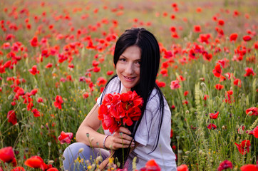 a young girl with dark long hair is wearing a blouse T-shirt and blue jeans sitting with a bouquet of poppy flowers among the poppy field