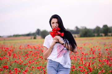 a young girl with dark long hair is wearing a blouse T-shirt and blue jeans standing among a poppy field