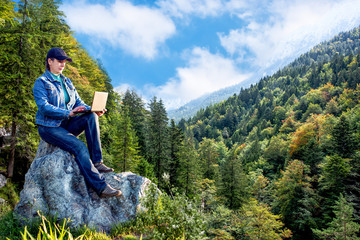 A girl sitting on rock with laptop notebook on background of mountains. Travel.
