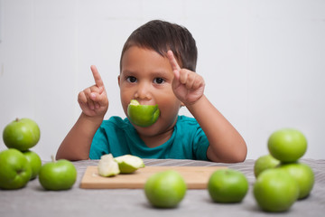 A cute young boy signaling with his hands that he loves eating healthy raw fruits in his diet, and holding a green apple in his mouth.