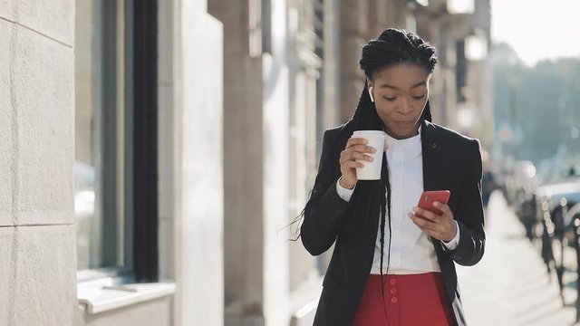 Portrait of a young African American businesswoman in a suit, walking around the city, drinking coffee and using smartphone. Concept: new business, communication, work day, freelancer.