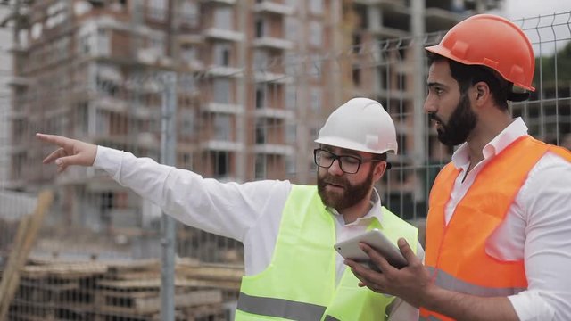 The builder and architect man are discussing the construction plan of the modern business center standing near construction site. They checks the work with a tablet.