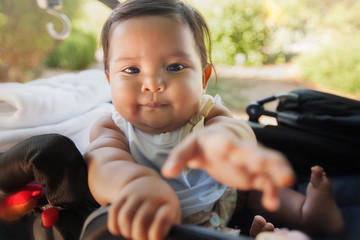 A cute baby using her hands to reach for something infront of her while riding on a carriage type of stroller.