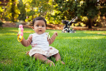 A happy hispanic baby playing and grasping a few chewable baby toys while sitting on the lawn of a park with he stroller in the background. - Powered by Adobe