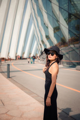 Naklejka premium Portrait of Beautiful Asian Woman in Fashionable Black Dress Clothing With Her Hat While Relaxing on Vacation, Beauty Woman Relax Posing in Fashion Clothes on Modern Building Background.