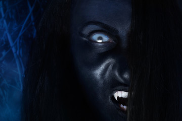 Creepy dark vampire face with white eyes and fangs on night wood background.