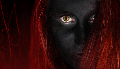 Face of a witch with black skin, cat eyes and red hair on burning wood background, close-up.