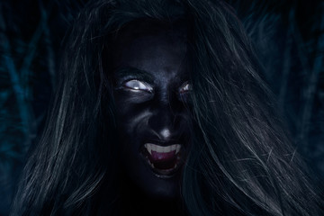 Scary face of silvan witch with dark skin and fangs screaming in night wood.