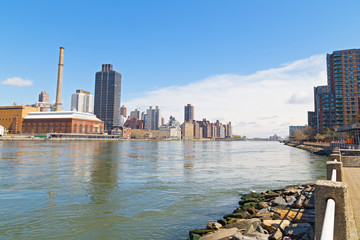 East River landscape with Manhattan on the left and Roosevelt Island apartments complex on the right. Urban landscape with river view and bright blue skies.