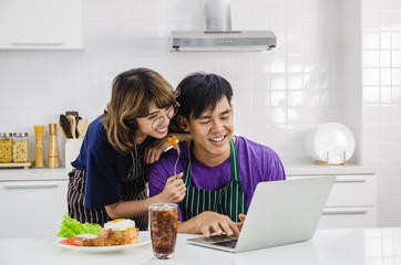 Smiling happy young Asian couple wearing apron and cooking together and feeding boyfriend while he is typing on laptop other in a white kitchen background.