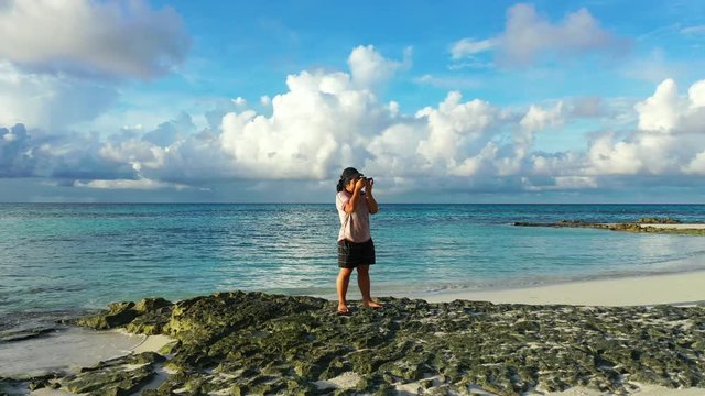 Young girl taking pictures with her camera in a small rocky beach in Thailand. Turquoise waters behind her