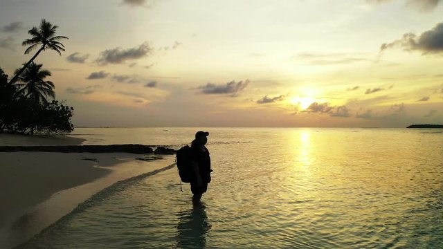 female traveler with a backpack and a cap standing in the water and enjoying the beautiful sunset in a beach in the Maldives Islands