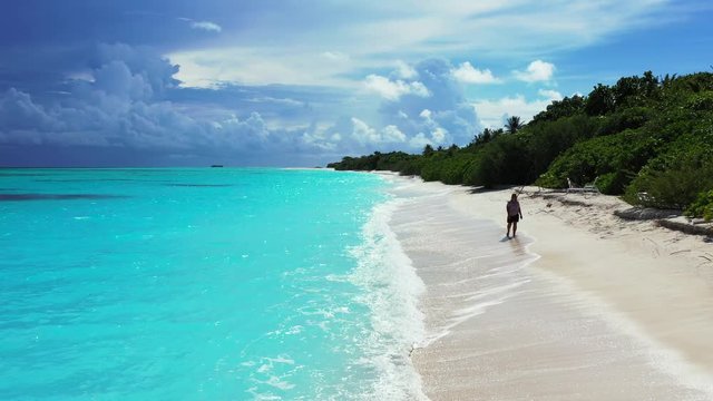 A woman walking along the beach through waves, facing the camera. On an island in the turquoise Caribbean Sea with a dramatic sky in the background. Wide angle, aerial dolly out.