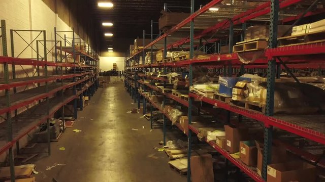 Aerial footage of interior of industrial warehouse. Drone films forward and slowly moves upward between half empty shelves in a long aisle.