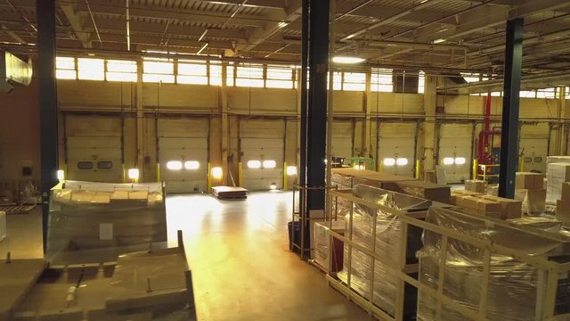 Aerial view of the inside of an industrial warehouse. Sun shines in on packed furniture through windows and bay doors, as drone circles and moves to the right.