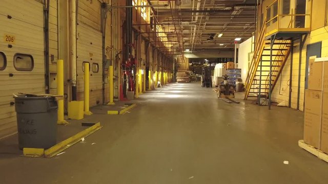 Interior Of Industrial Warehouse. Drone Moves Slowly Forward Low Above The Floor. Passes By Bay Doors On The Left And Stacked Goods And Equipment On The Right.