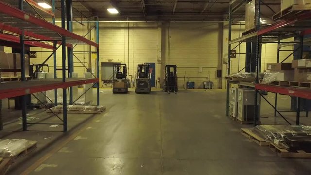 Aerial view of half empty shelves in an industrial warehouse. Drone flies slowly forward between the shelves toward wall and small forklifts.