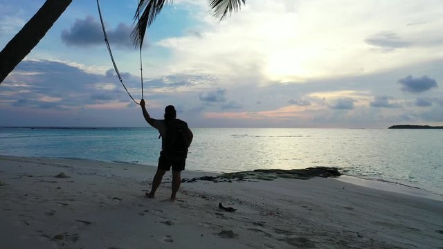 Trucking shot of female tourist with luggage holding rope hanging from tree on the sandy beach with palm trees waiting for sunrise, night sky and silhouettes