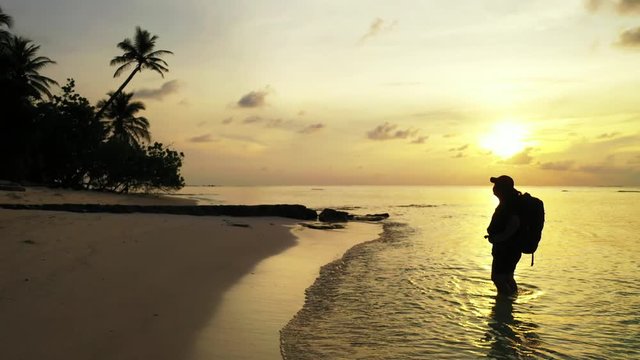 A silhouette of a man wearing a backpack and a cap is staying in shallow water while looking onto a Caribbean island. Iit by the golden light of the setting sun. Wide angle, truck left