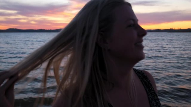 Handheld, slow motion shot of a woman touching her head and smiles, at the beach, at lake Huma, sunset, on a sunny evening, at Hume Reservoir, at New South Wales and Victoria, in Australia