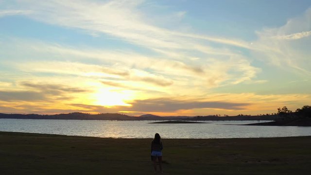Aerial, drone shot panning around a woman, standing on a beach, at lake Huma, sunset, on a sunny evening, at Hume Reservoir, between New South Wales and Victoria, in Australia